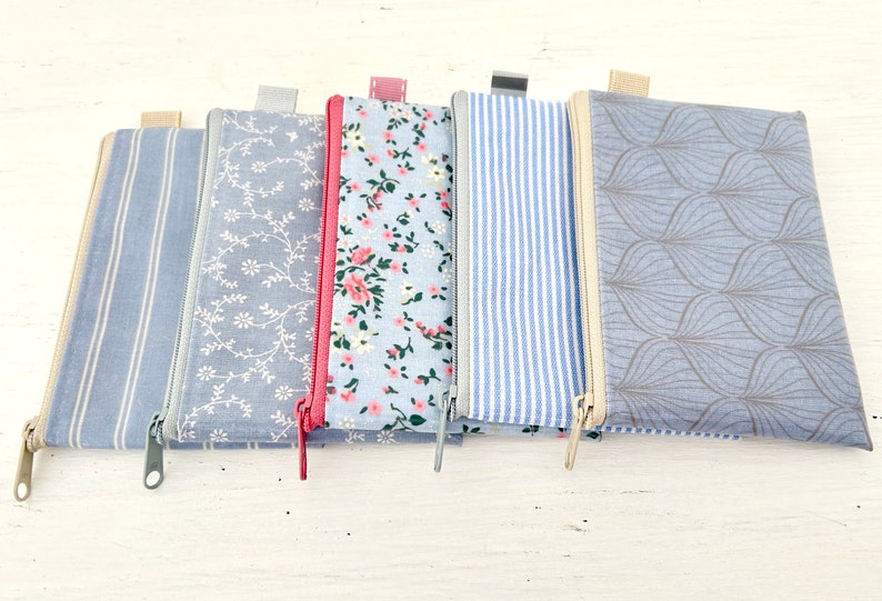 May include: Five zippered pouches with different patterns. The pouches are all made of a light blue fabric with a white pattern. The patterns include stripes, floral, and geometric designs.