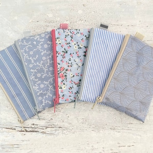 May include: Five zippered pouches with different patterns. The pouches are made of a light blue fabric with white designs. The patterns include stripes, floral, and a leaf design. The pouches are arranged in a fan shape.