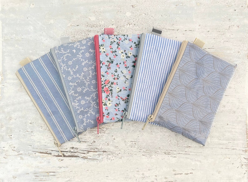 May include: Five zippered pouches with different patterns. The pouches are made of a light blue fabric with white designs. The patterns include stripes, floral, and a leaf design. The pouches are arranged in a fan shape.
