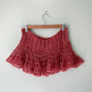 May include: A crocheted shawl in a dusty rose color, hanging on a wooden hanger against a light green wall.