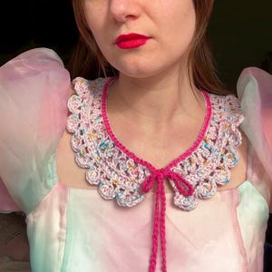 May include: A white crochet collar with pink trim and a pink bow tie. The collar is decorated with colorful flecks.