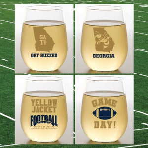 May include: Four clear wine glasses with gold lettering and graphics. The glasses feature the following designs: "GA TECH GET BUZZED", "GEORGIA" with a bee graphic, "YELLOW JACKET FOOTBALL", and "GAME DAY!" with a football graphic.