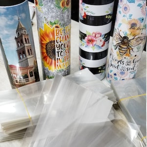 May include: A stack of clear plastic bags, likely for packaging, are on a white surface.  There are four tumblers in the background, each with a different design.  One tumbler has a sunflower design with the text "Be the change you want to see in the world".  Another tumbler has a black and white striped design with floral accents.  A third tumbler has a church building with a white steeple.  The final tumbler has a bee and sunflower design with the text "Words are honey sweet to the soul".