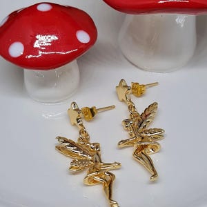 May include: Gold fairy dangle earrings with a star charm. The fairies are wearing a dress and have wings. The earrings are on a white plate with a red mushroom.