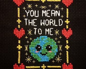 May include: Cross-stitch embroidery on black fabric with a yellow and red border. The embroidery depicts a green and blue earth with a smiling face and the text "You mean the world to me".