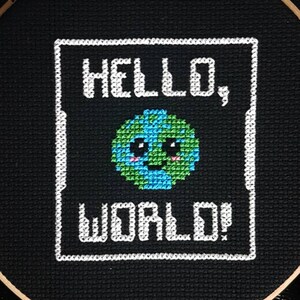 May include: Embroidered hoop art with a black background and white text that reads "HELLO, WORLD!" with a cartoon earth with a smiling face in the center.