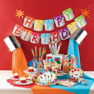 May include: A colorful paint can themed birthday party with a banner that says "Happy Birthday". The table is set with paint splattered paper plates, cups, and napkins. There are also paintbrush shaped lollipops and cupcakes.