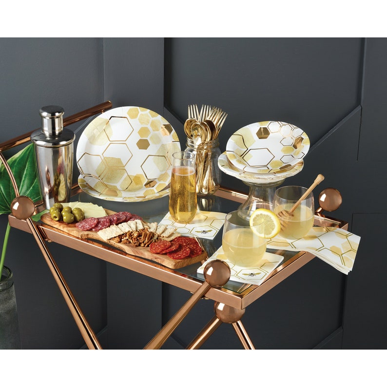 May include: A gold and white honeycomb patterned paper party plate set with a silver cocktail shaker, a wooden serving tray with salami, olives, and crackers, two glasses of lemonade, and gold and white honeycomb patterned napkins.