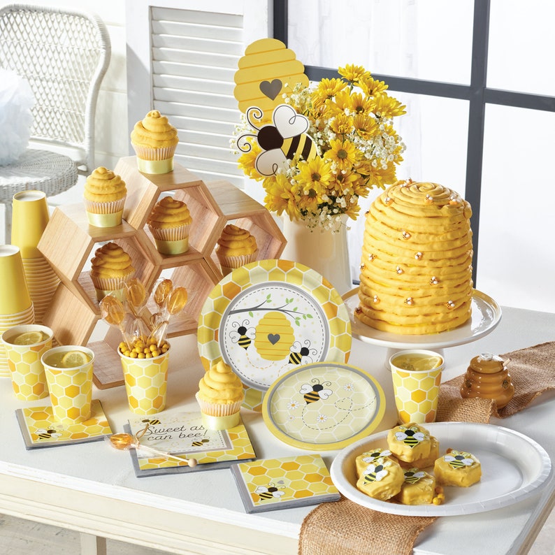 May include: A yellow and black bee-themed baby shower party table setting with cupcakes, a beehive cake, and paper plates and cups with a beehive pattern. The table is decorated with a burlap runner and a yellow and black beehive centerpiece.