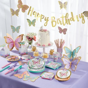 May include: A purple table setting with butterfly themed party supplies. The table includes plates, napkins, cups, straws, and a cake. The centerpiece is a white cake with a butterfly on top. The table is decorated with a gold "Happy Birthday" banner and butterfly decorations.
