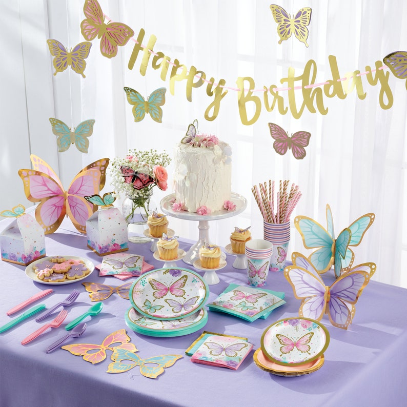May include: A purple table setting with butterfly themed party supplies. The table includes plates, napkins, cups, straws, and a cake. The centerpiece is a white cake with a butterfly on top. The table is decorated with a gold "Happy Birthday" banner and butterfly decorations.