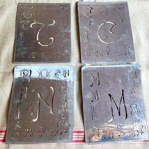 Könnte beinhalten: Vier antike Metall-Schablonen mit verschiedenen Buchstaben- und Designmustern. Die Schablonen sind aus Metall und haben einen abgenutzten, antiken Look. Die Schablonen sind in zwei Reihen zu je zwei angeordnet. Die obere Reihe zeigt Schablonen mit den Buchstaben "C" und "G" und die untere Reihe zeigt Schablonen mit den Buchstaben "R" und "M".