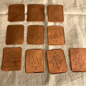 Könnte beinhalten: Zehn Kupferschablonen mit filigranen Designs für Stickereien. Jede Schablone zeigt ein anderes Monogramm mit den Buchstaben 'G' und 'M' und einer Nummer. Die Schablonen sind rechteckig und haben abgerundete Ecken.