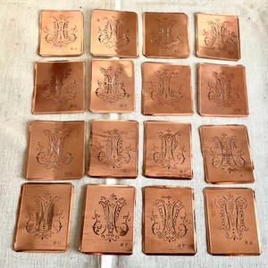 以下が含まれることがあります： 装飾的なMの文字のデザインが施された銅製のステンシル16枚セット。各ステンシルには、「M.A.」や「M.B.」などの文字の組み合わせがラベル付けされています