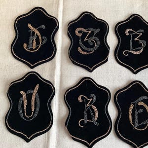 May include: Six black velvet coasters with gold and black beaded embroidery of the letters J, G, L, U, K, and D.