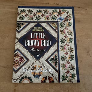 Könnte beinhalten: Ein Buch mit dem Titel "Little Brown Bird Patterns" mit einem dekorativen floralen und geometrischen Design. Das Cover zeigt den Text "Applique Masterpiece" und den Namen des Autors, Margaret Docherty. Das Buch hat einen dunkelblauen Rand.
