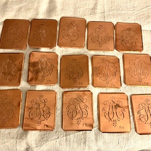Könnte beinhalten: Eine Sammlung kupferfarbener rechteckiger Schablonen mit verzierten, stilisierten Buchstabenmustern. Jede Schablone zeigt ein anderes Monogramm, wobei die Buchstaben ausgeschnitten sind. Die Schablonen sind auf einer weißen Stoffoberfläche angeordnet.