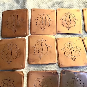 May include: A selection of vintage stencils featuring elaborate letter designs. Each stencil is a square shape with a cut-out letter, surrounded by decorative flourishes. The stencils are a warm brown colour, with some showing signs of age and wear.