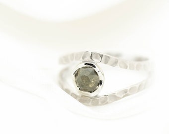 May include: A silver ring with a hammered texture and a central, faceted, gray gemstone. The ring's design features a split band that curves around the stone, creating a modern and elegant look. The ring is set against a white background.