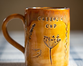 May include: A ceramic mug with a brown and yellow glaze. The mug features the text "Culley's Cup" and etched botanical designs. The handle is a matching brown color. The mug is sitting on a blue and white checkered surface.