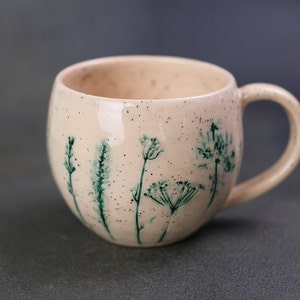 May include: A speckled, cream-colored ceramic mug with a rounded shape and a handle. The mug is decorated with hand-painted green botanical designs. The interior of the mug is visible. The mug is suitable for coffee, tea, or other beverages.