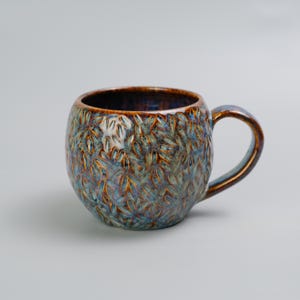 Può includere: Una tazza in ceramica con superficie strutturata e forma arrotondata. La tazza presenta un motivo di disegni a forma di foglia nei toni del blu, marrone e oro. Ha un interno marrone scuro e un manico abbinato.