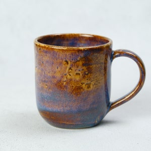 Può includere: Una tazza in ceramica con base arrotondata e manico curvo. La tazza ha una finitura lucida con una miscela di tonalità marroni, dorate e blu. L'interno della tazza è di una tonalità di blu più scura.