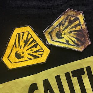 May include: Two yellow and black hazard patches with an explosion graphic. The patches are hexagonal and have a black border. A yellow caution tape is visible at the bottom of the image.
