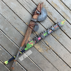 Könnte beinhalten: Zwei handgefertigte Holz-Waffen. Eine hat zwei Kreissägeblätter, die mit roter Schnur an einem Holzgriff befestigt sind. Die andere hat Metallspitzen und bunte Farbe. Der Text "RAAPTORGEDDON" ist sichtbar.