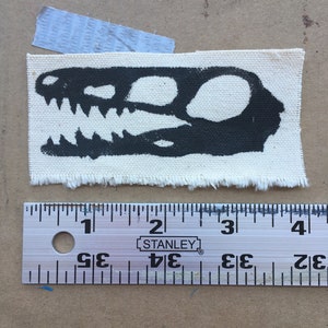 Op de afbeelding: Een zwart dinosaurus schedel ontwerp is gedrukt op een klein, rechthoekig stukje off-white stof. De stof heeft een gerafelde rand. Een metalen liniaal is eronder geplaatst.