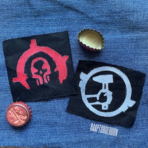 May include: Two black fabric patches with white and red designs, and two bottle caps. One patch features a red skull design, and the other has a white hammer and hand design. The text "RAAPTORGEDDON" is at the bottom.