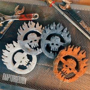 May include: Four metal skull emblems in various colors: white, silver, gray, and orange. Each emblem features a skull within a flaming circle design. The word "RAAPTORGEDDON" is printed in white at the bottom of the image. Tools are in the background.
