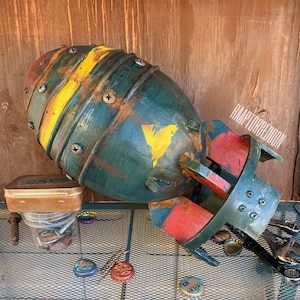 May include: A handmade, weathered-look bomb prop in shades of green, yellow, and red, with visible screws and a distressed finish. The prop is lying on a metal surface, with bottle caps and a small container nearby. The text "RAAPTORGEDDON" is visible.