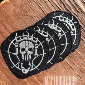 Puede incluir: Un conjunto de cinco parches negros y circulares con un diseño de calavera blanca y el texto "RAAPTORGEDDON". La calavera tiene una corona y está rodeada por un diseño circular blanco.