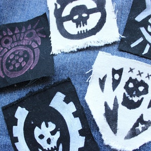 Può includere: Una collezione di toppe in tessuto con vari disegni disegnati a mano. Le toppe presentano motivi di teschi e ingranaggi in bianco e nero e un disegno viola su una toppa nera. Le toppe sono posizionate su uno sfondo di denim blu.
