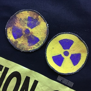 Op de afbeelding: Twee ronde patches met het paarse stralingssymbool op een gele achtergrond. De patches hebben een verweerde uitstraling. Ze zijn geplaatst op een donkerblauwe stof.