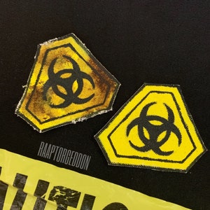 Op de afbeelding: Twee gele en zwarte biohazard patches met een verweerde uitstraling. Eén patch heeft een bruine, verouderde uitstraling. De patches liggen op een zwarte achtergrond. Onderaan een gele waarschuwingstape met de tekst "RAAPTORGEDDON".