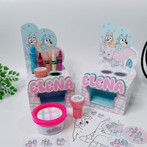 Puede incluir: Un personaje de dibujos animados de perro azul y marrón con un elefante rosa en una caja con estampado azul y blanco con el nombre "Elena" en ella. La caja contiene lápices de colores, plastilina y un sello. También hay pegatinas y una página para colorear con el mismo personaje de dibujos animados de perro.