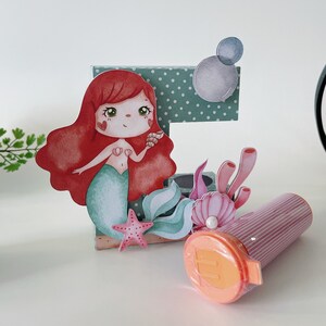Puede incluir: Un expositor decorativo con una sirena de dibujos animados de pelo largo rojo y cola verde azulado, situada frente a una letra verde con lunares. Incluye una estrella de mar rosa, coral, una concha con una perla y un recipiente cilíndrico de rayas rosas.