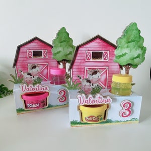 Puede incluir: Dos cajas de color rosa y blanco con forma de granero con un diseño de vaca y ternero. Cada caja tiene un recipiente rosa y amarillo con el nombre "Valentina" y el número "3" en la parte delantera. Las cajas contienen Play-Doh y espuma.