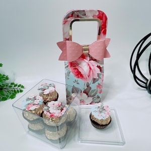 May include: A clear plastic box filled with chocolate candies wrapped in gold foil and topped with floral paper designs. The box is next to a floral patterned paper gift bag with a pink bow and a clear plastic tray with a single chocolate candy.