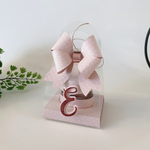 Peut inclure: Une boîte cadeau rose à pois blancs avec une fenêtre en plastique transparent et un nœud rose. La boîte a une poignée et la lettre "E" est sur le devant.