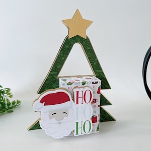 Puede incluir: Un árbol de Navidad verde con una estrella dorada en la parte superior. Una cara de Papá Noel con un gorro rojo está unida al frente del árbol. El árbol está decorado con una caja blanca que dice "HO HO HO" en letras verdes.