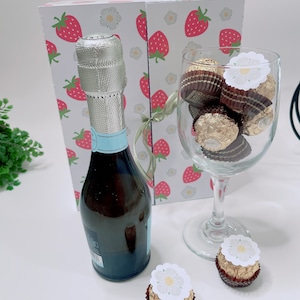 Puede incluir: Un set de regalo con una botella de vino espumoso, una copa de vino llena de bombones y dos bombones individuales. El fondo presenta una caja decorada con fresas y flores blancas.