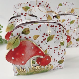 Puede incluir: Una caja de regalo blanca con un diseño de hongo rojo y blanco. La caja tiene un asa dorada y está decorada con bayas rojas y hojas verdes. La caja es perfecta para guardar pequeños regalos o favores.