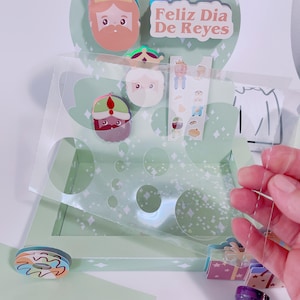 Puede incluir: Una caja de manualidades de papel verde y blanca con una tapa de plástico transparente. La caja tiene un diseño festivo con una estrella, una corona y las palabras "Feliz Dia De Reyes". La caja está llena de pequeñas pegatinas de colores y una almohadilla de tinta morada.