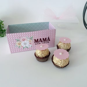 May include: A pink and grey gift box with the words "MAMÁ amor infinito" printed on the front. The box is filled with three chocolate truffles, each topped with a pink flower.