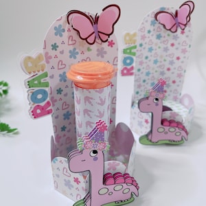 May include: A whimsical party favor set with a pink dinosaur theme. The set includes decorative boxes with floral and heart patterns, dinosaur cutouts, and butterfly accents. The word "ROAR" is displayed on the boxes. A clear tube with an orange lid is also included.