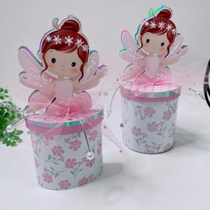 May include: Two cylindrical containers featuring a white and pink floral pattern. Each lid is topped with a cutout of a fairy with red hair, a flower crown, and pink wings. The fairy figures are accented with pink tulle and white pearl beads.