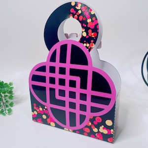 Puede incluir: Una caja de regalo decorativa con fondo negro y diseño geométrico rosa. La caja tiene asa y está decorada con puntos rosas, rojos y amarillos tipo confeti. La caja está sobre una superficie blanca.